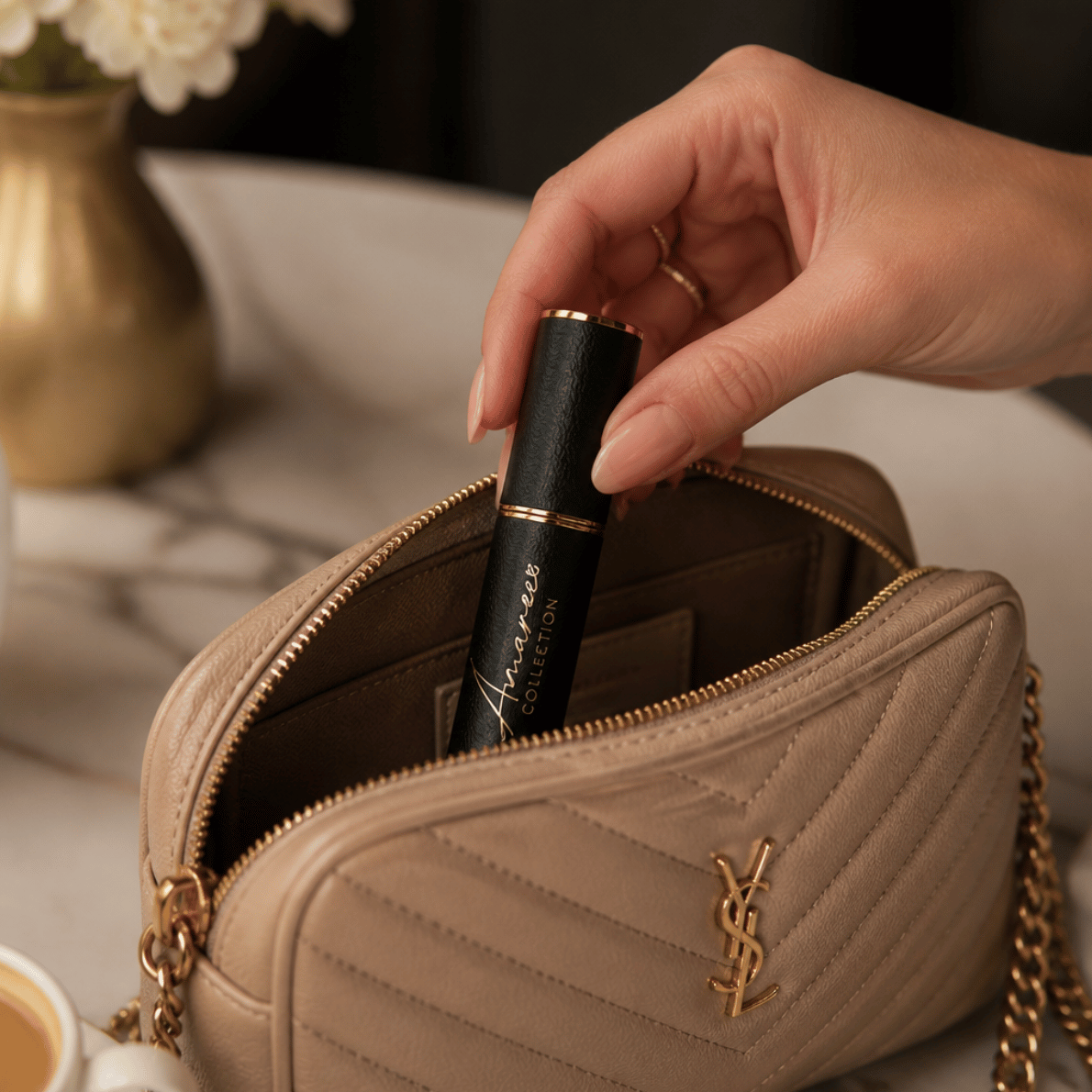 A beige quilted cosmetic bag with gold-tone hardware and a YSL monogram logo, shown open with a hand inserting a black and gold tube into the interior. The scene is styled on a light marble surface with a blurred gold vase and white flowers in the background.