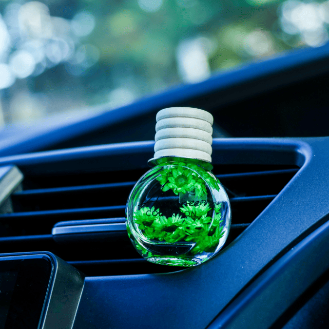 A small clear glass car air freshener bottle with a white ribbed cap, filled with bright green botanical pieces, resting on a dark car dashboard vent.