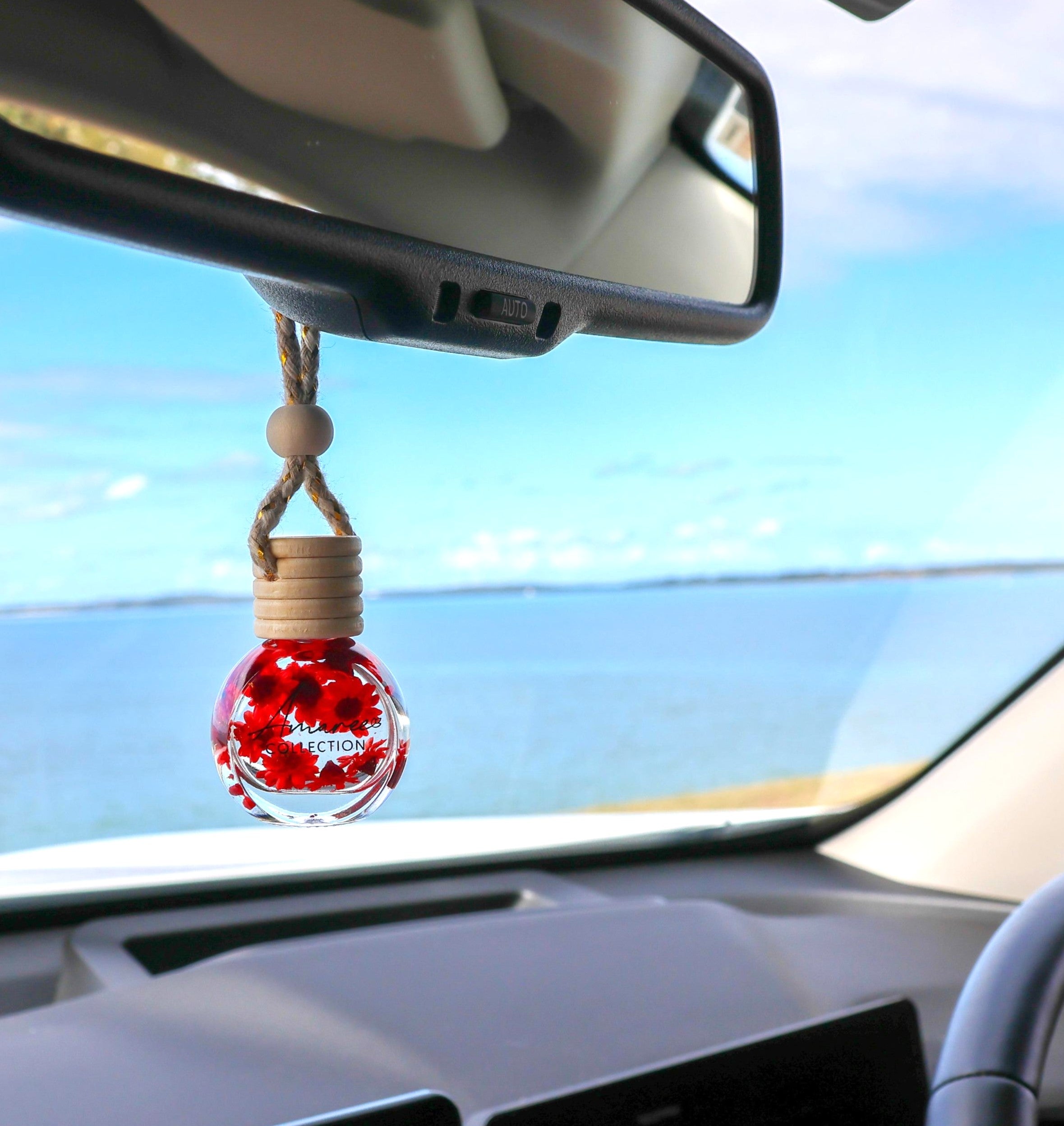 A car air freshener hanging ornament inside a vehicle, featuring a clear glass bottle filled with red maple leaves and a wooden cap, suspended from a rearview mirror.