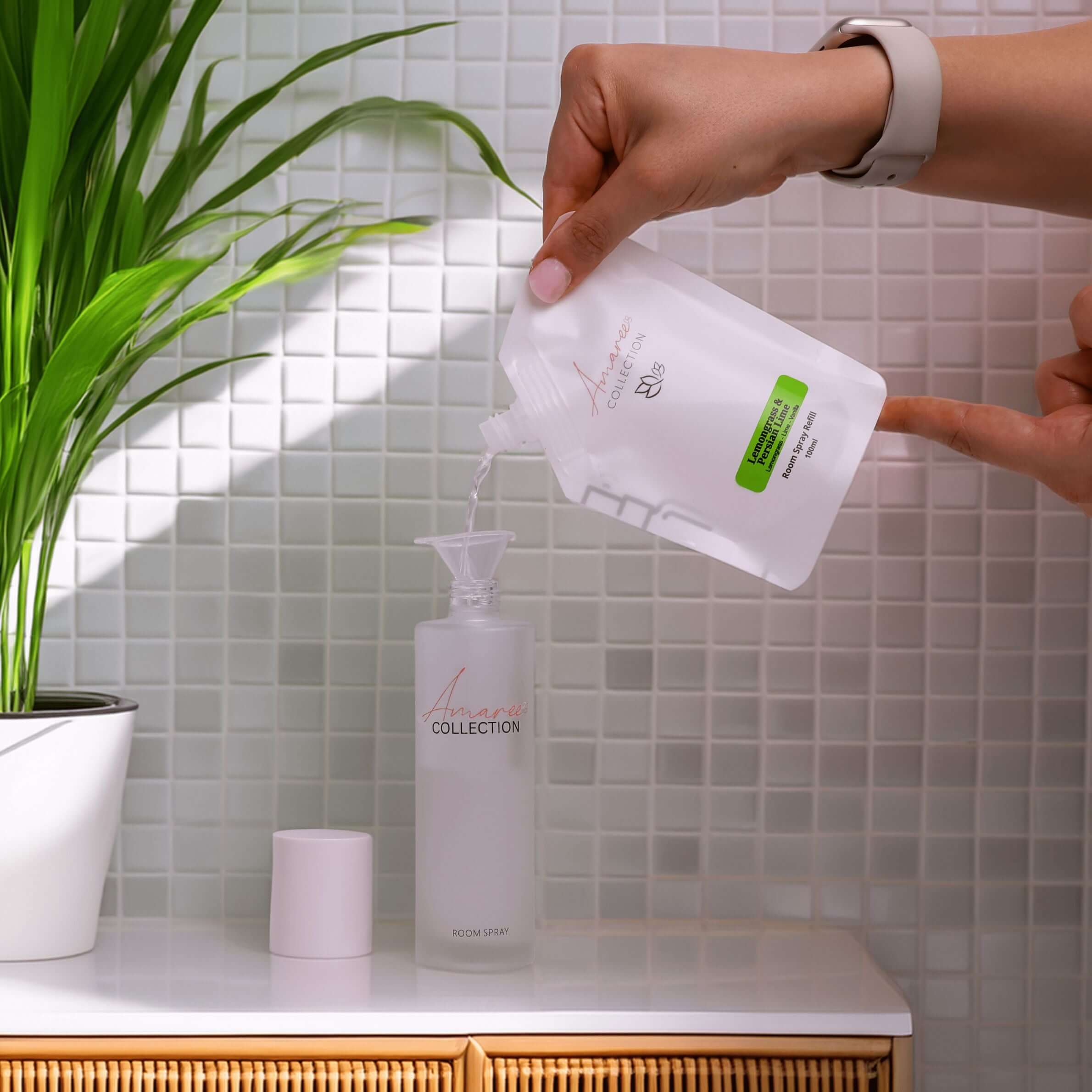 Person pouring Lemongrass Room Spray Refill 100ml into a clear spray bottle next to a green plant.