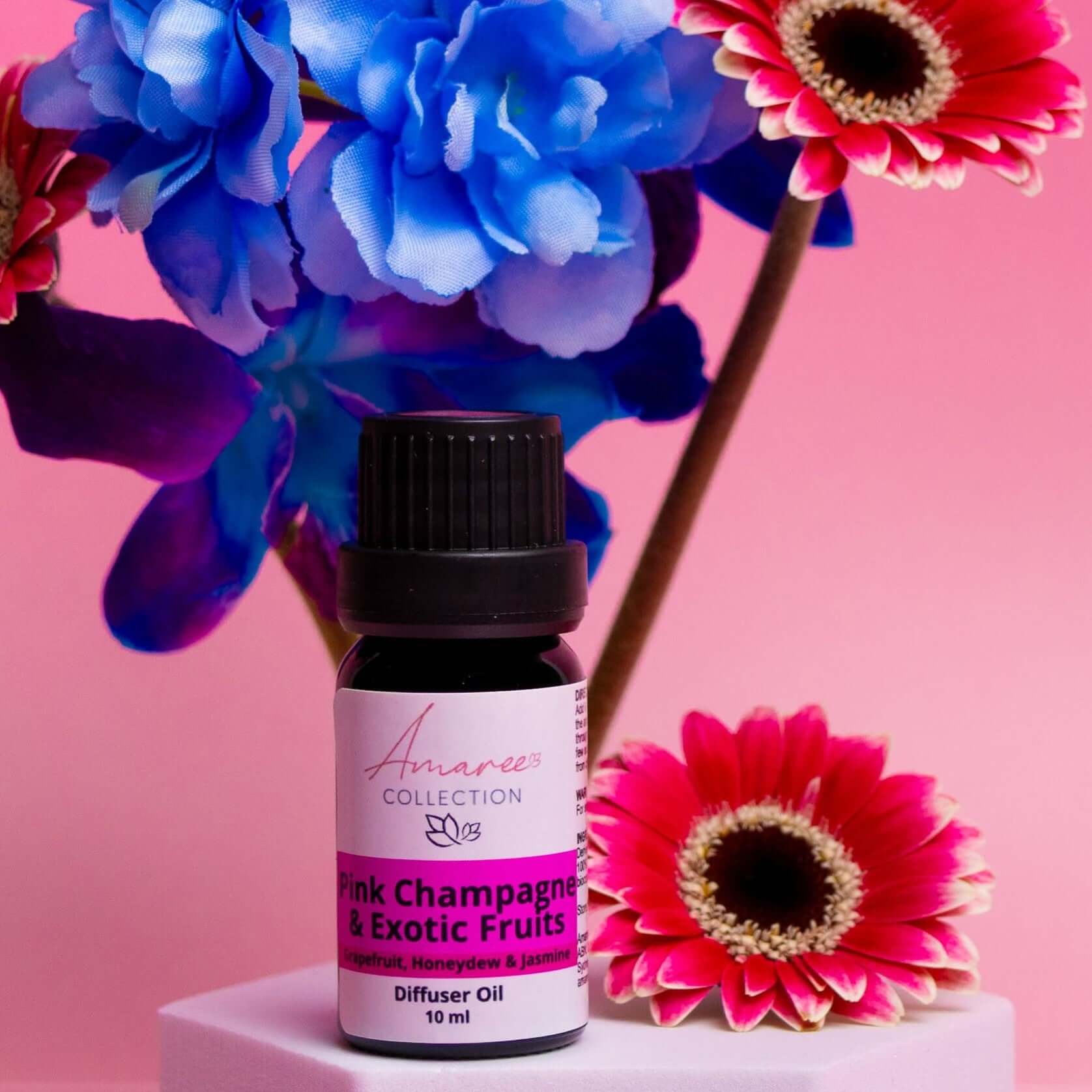 Pink Champagne & Exotic Fruits Diffuser Oil bottle surrounded by colorful flowers on a pink background.