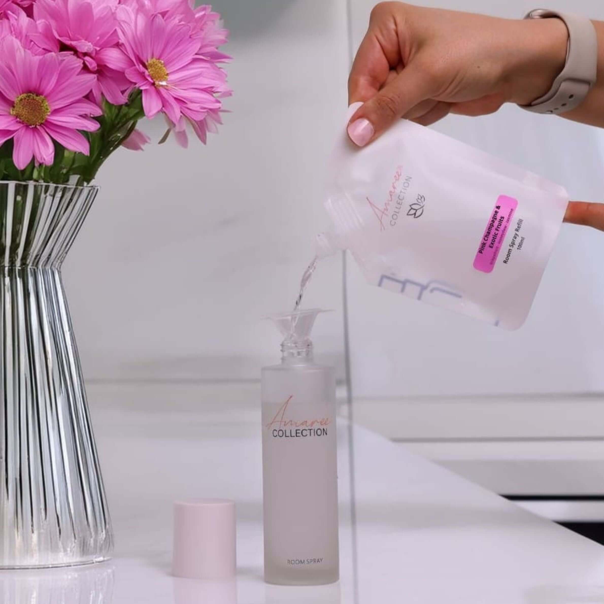 Pouring Pink Champagne & Exotic Fruits Room Spray Refill 100ml into spray bottle next to flowers.