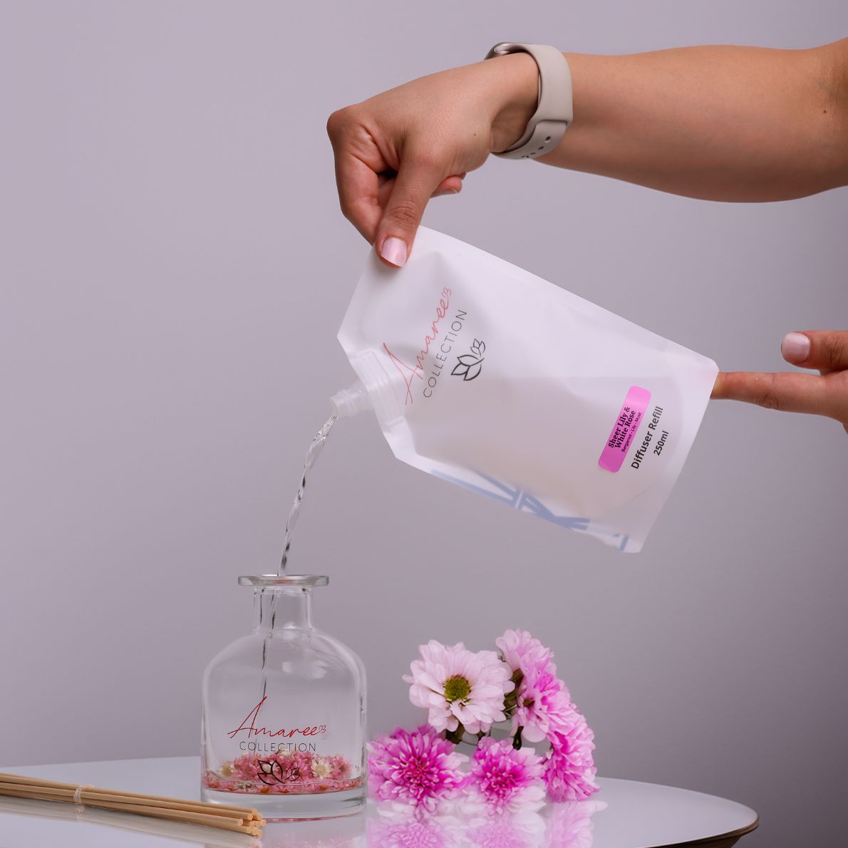 Hand pouring a liquid from a white pouch into a glass bottle with pink flowers on a gray background
