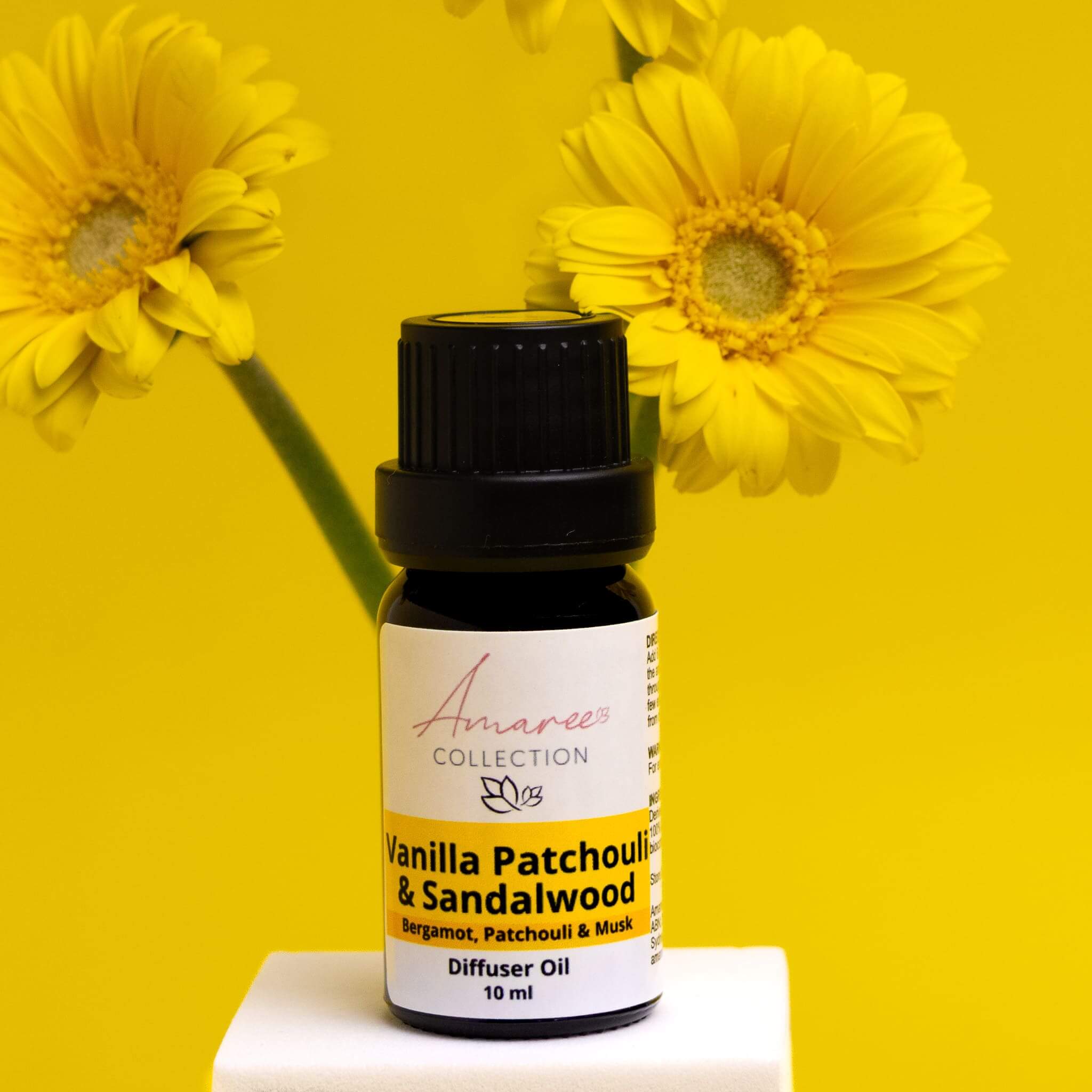 Vanilla Patchouli & Sandalwood Diffuser Oil bottle against a bright yellow backdrop with flowers.