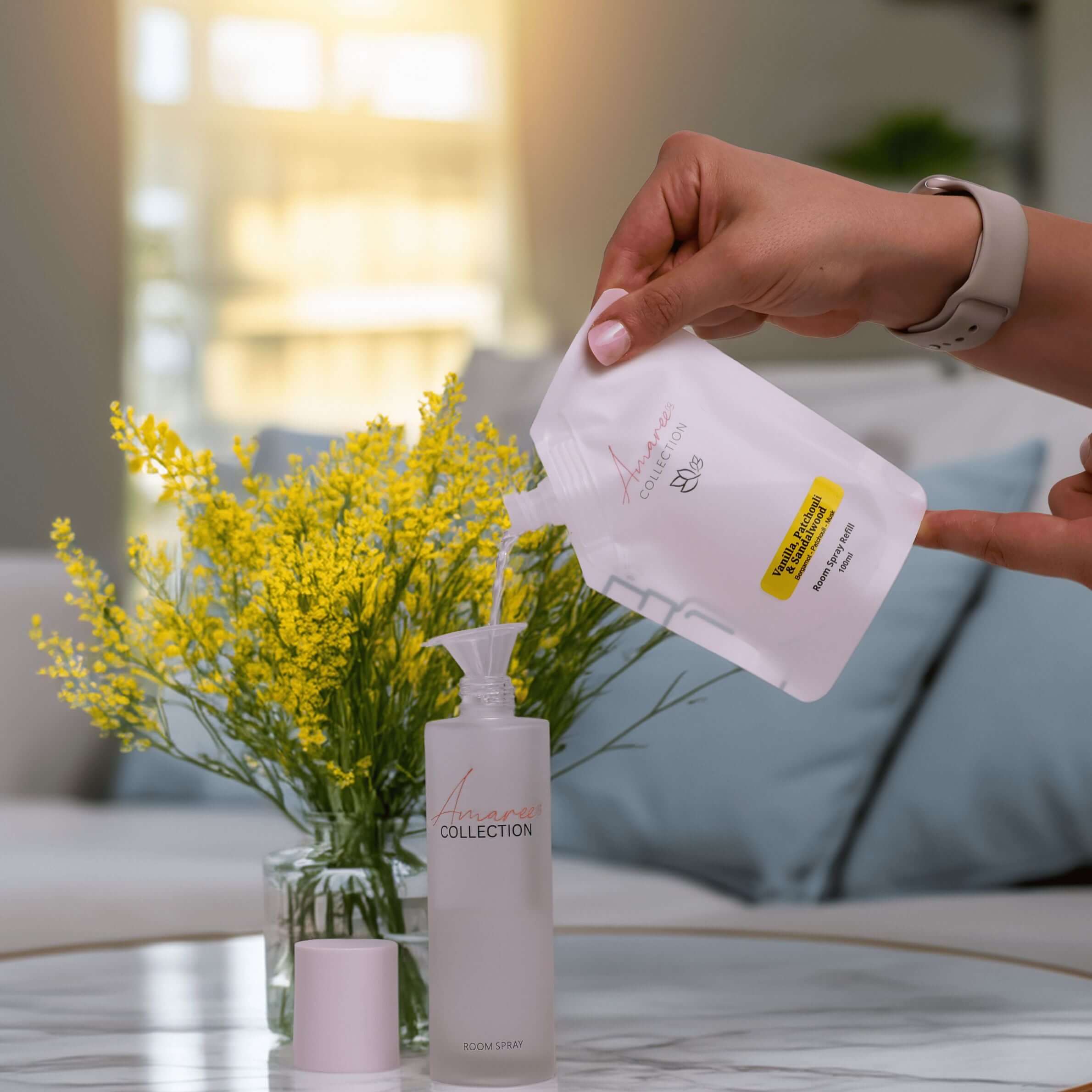 Hand pouring Vanilla Patchouli & Sandalwood Room Spray Refill 100ml into a clear bottle beside yellow flowers.