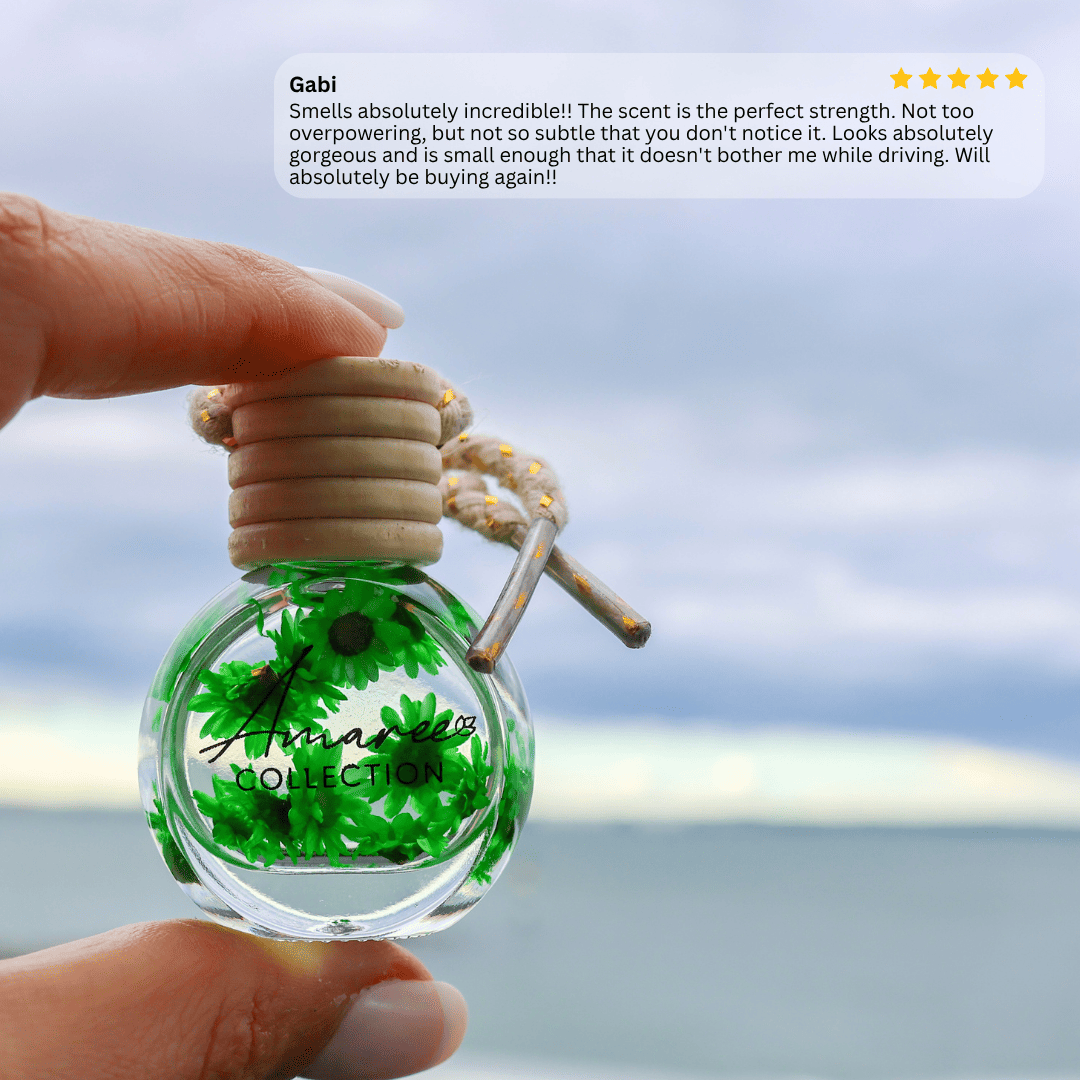 A hand holds a small clear glass perfume bottle with bright green botanical pieces inside and a light wood cap, against an outdoor blurred background of sky and water. A text box at the top reads &