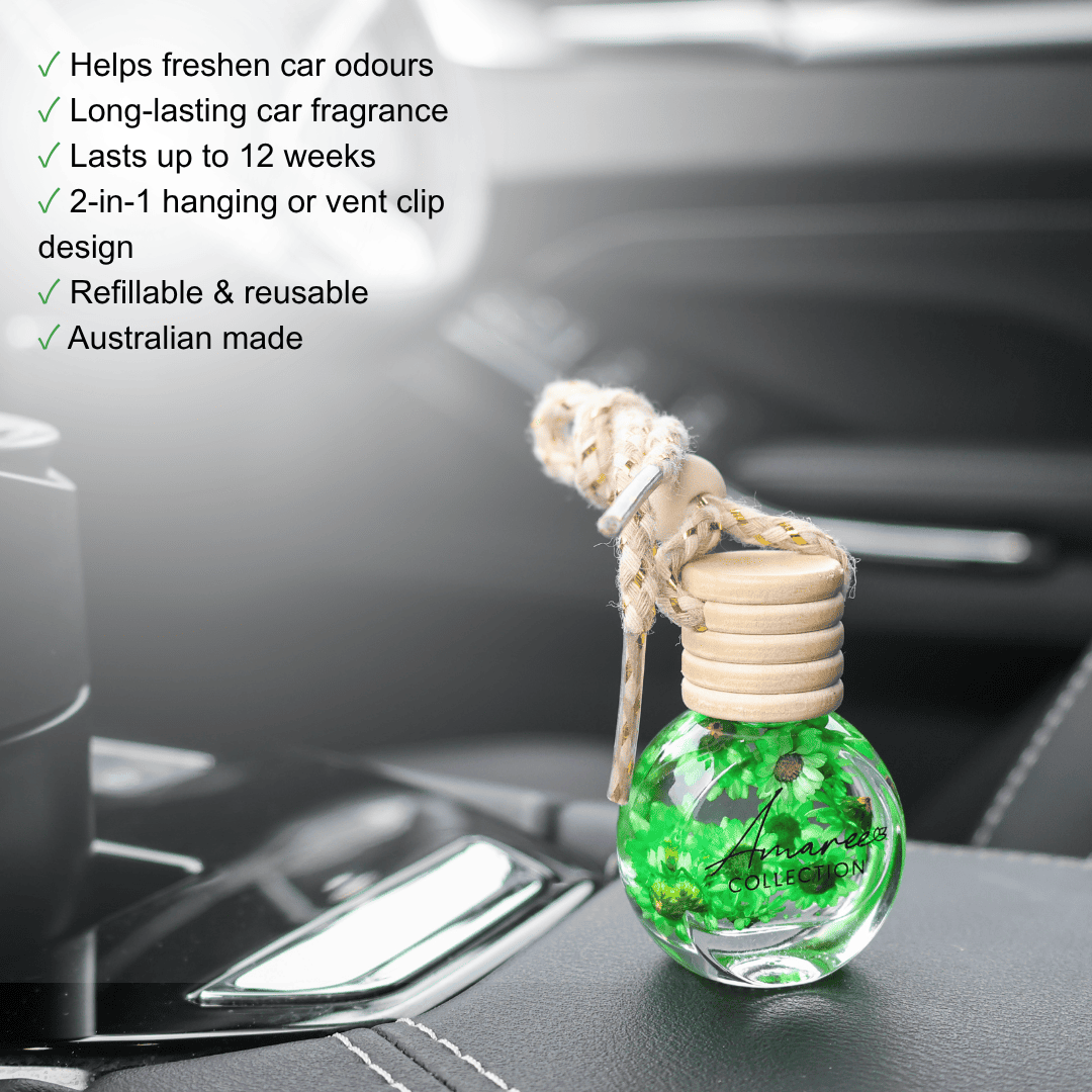 A small clear glass car fragrance bottle with bright green liquid and floating botanical pieces, hanging from a beige cord with a wooden cap, placed on a car dashboard. A list of product features appears on the left side.