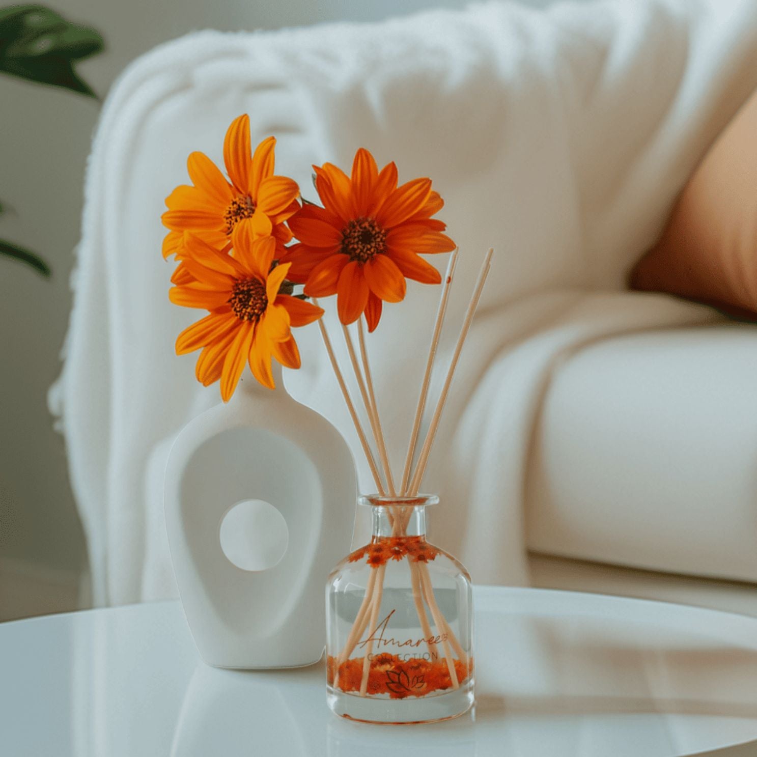Clear vase with orange flowers and sticks on a white surface with a blurred background