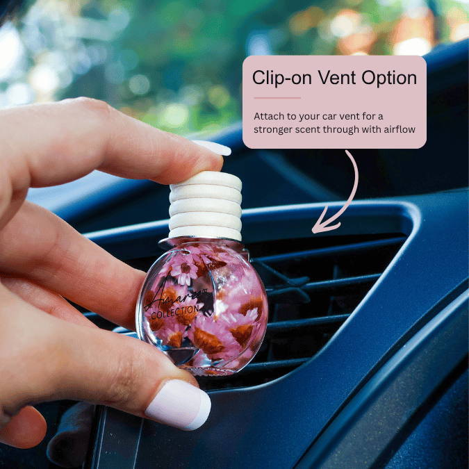 A hand holds a small round car air freshener bottle with pink floral contents and a white clip-on cap, positioned near a car vent. A pink text box reads &