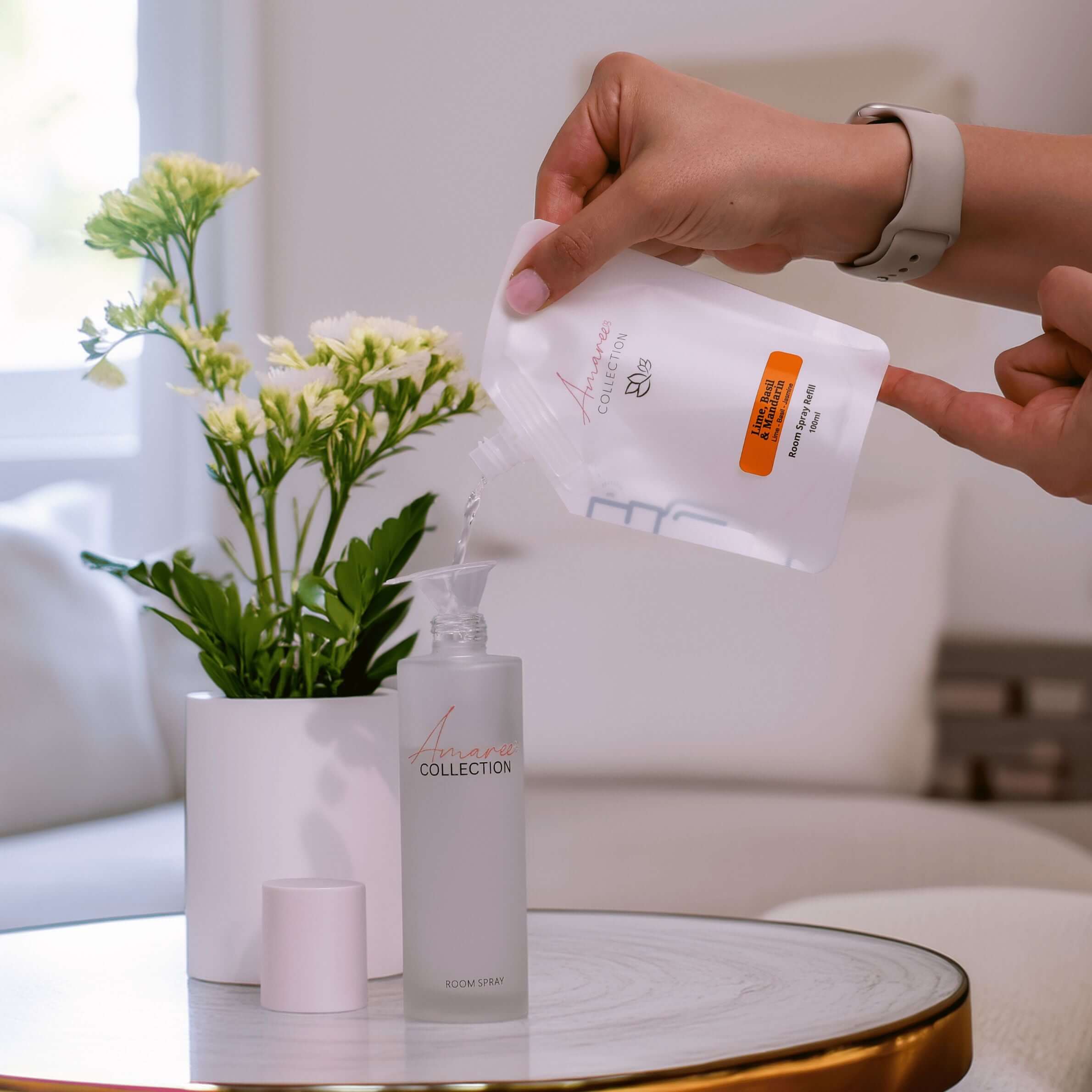 Lime Basil & Mandarin Room Spray Refill 100ml being poured into a spray bottle next to fresh flowers.