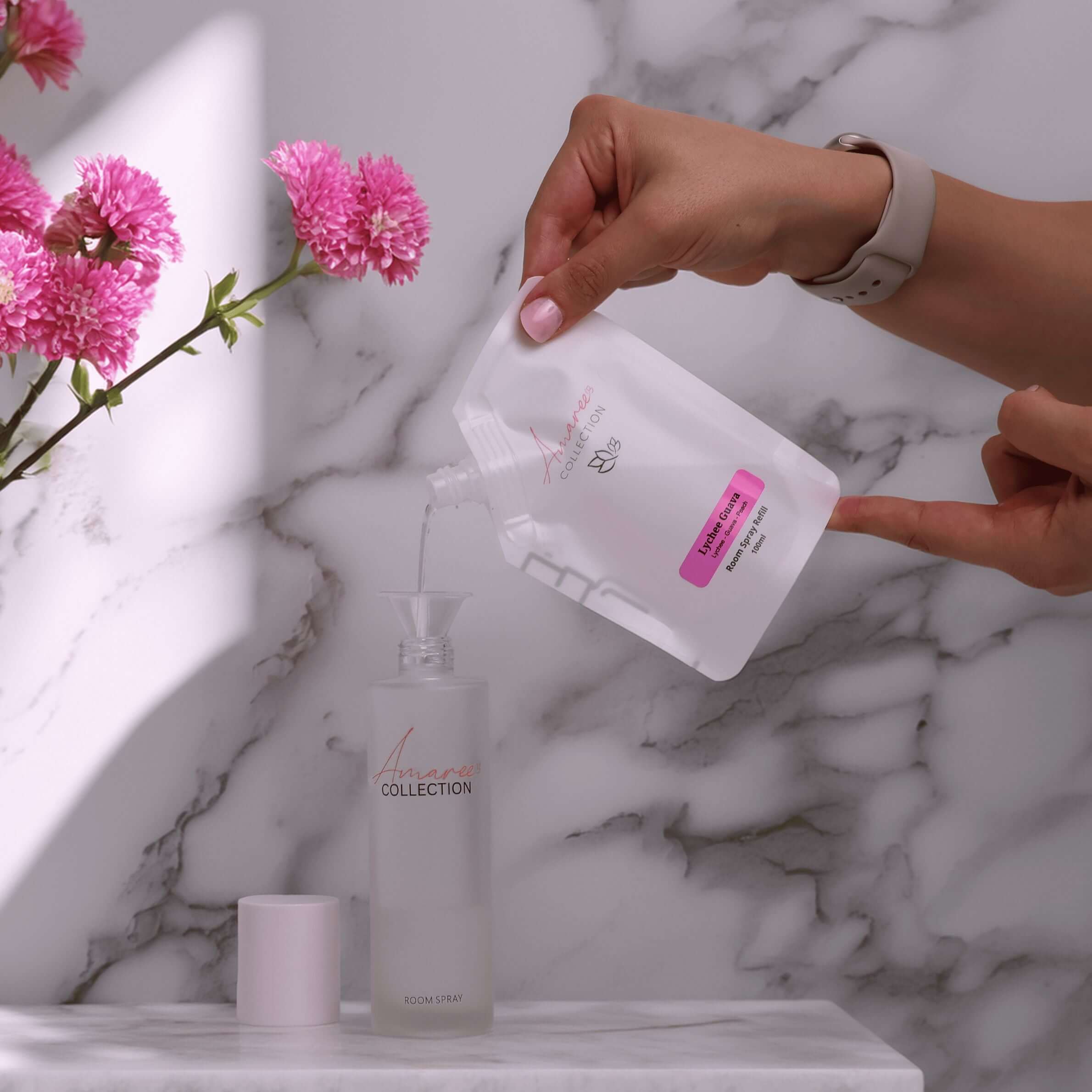 Pouring Lychee Guava Room Spray Refill 100ml into a bottle, surrounded by pink flowers on a marble countertop.