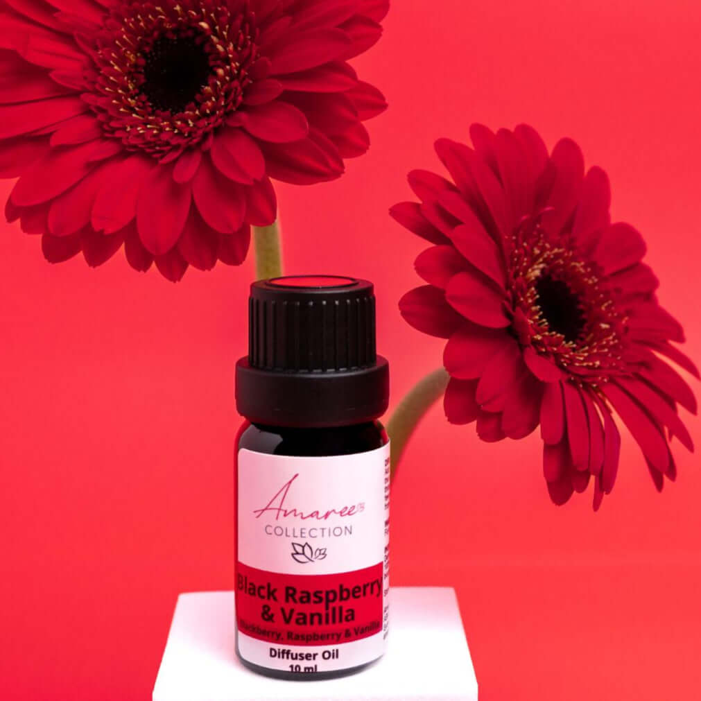 Black Raspberry & Vanilla Diffuser Oil bottle next to vibrant red gerbera daisies on a pink background.