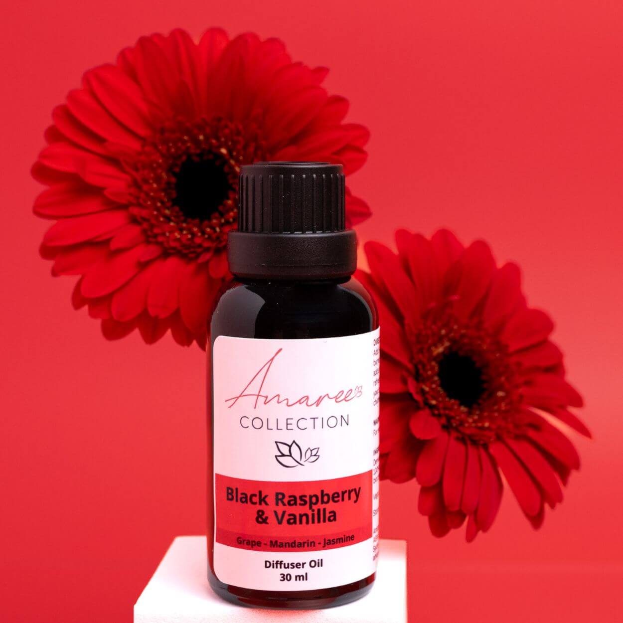 Black Raspberry & Vanilla Diffuser Oil bottle surrounded by vibrant red flowers on a red background.