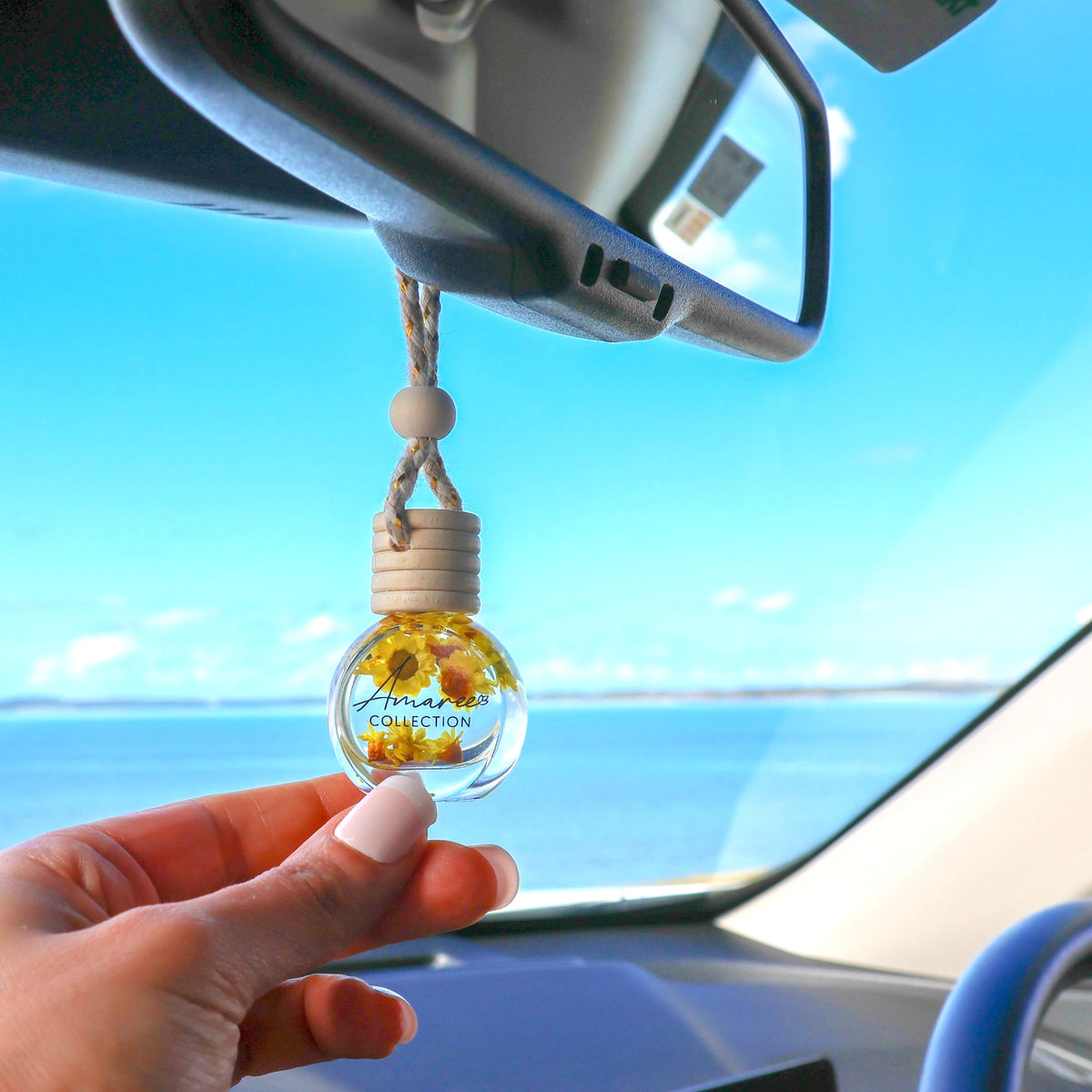 Car air freshener with sunflower design hanging from a car's rearview mirror against a blue sky.