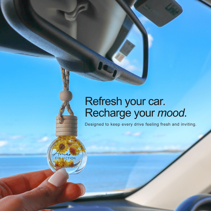 A hand holds a clear glass car air freshener bottle with yellow flower pieces inside, hanging from a car rearview mirror. The scene is set inside a car with a blue sky and ocean view outside, with text that reads &