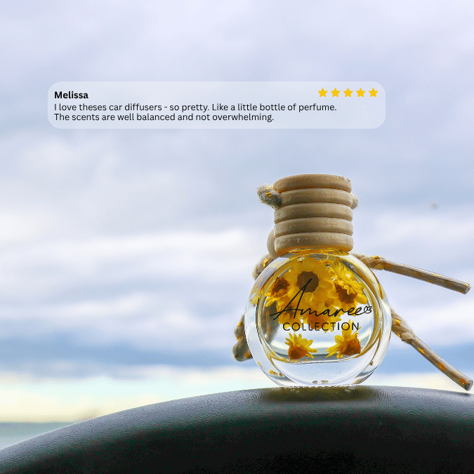 A small clear glass car diffuser bottle with yellow liquid and dried flower pieces inside, tied with a wooden loop, shown outdoors on a dark surface with a cloudy sky behind it. A review card for &