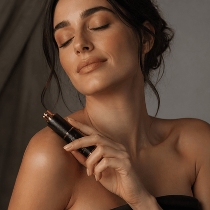 A woman with closed eyes holds a black cosmetic tube with a metallic rose gold cap against her shoulder, set against a dark gray background.