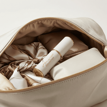 An open beige cosmetic bag with a gold zipper, shown filled with matching beige and white cosmetics and a satin scarf. The lipstick tube is branded &