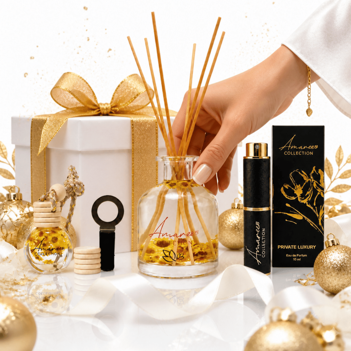 A hand holds a clear glass reed diffuser bottle with wooden reeds, surrounded by gold holiday ornaments, a white gift box with a gold ribbon, a small gold perfume bottle, and a black-and-gold Amaryllis Collection Private Luxury box on a white surface.