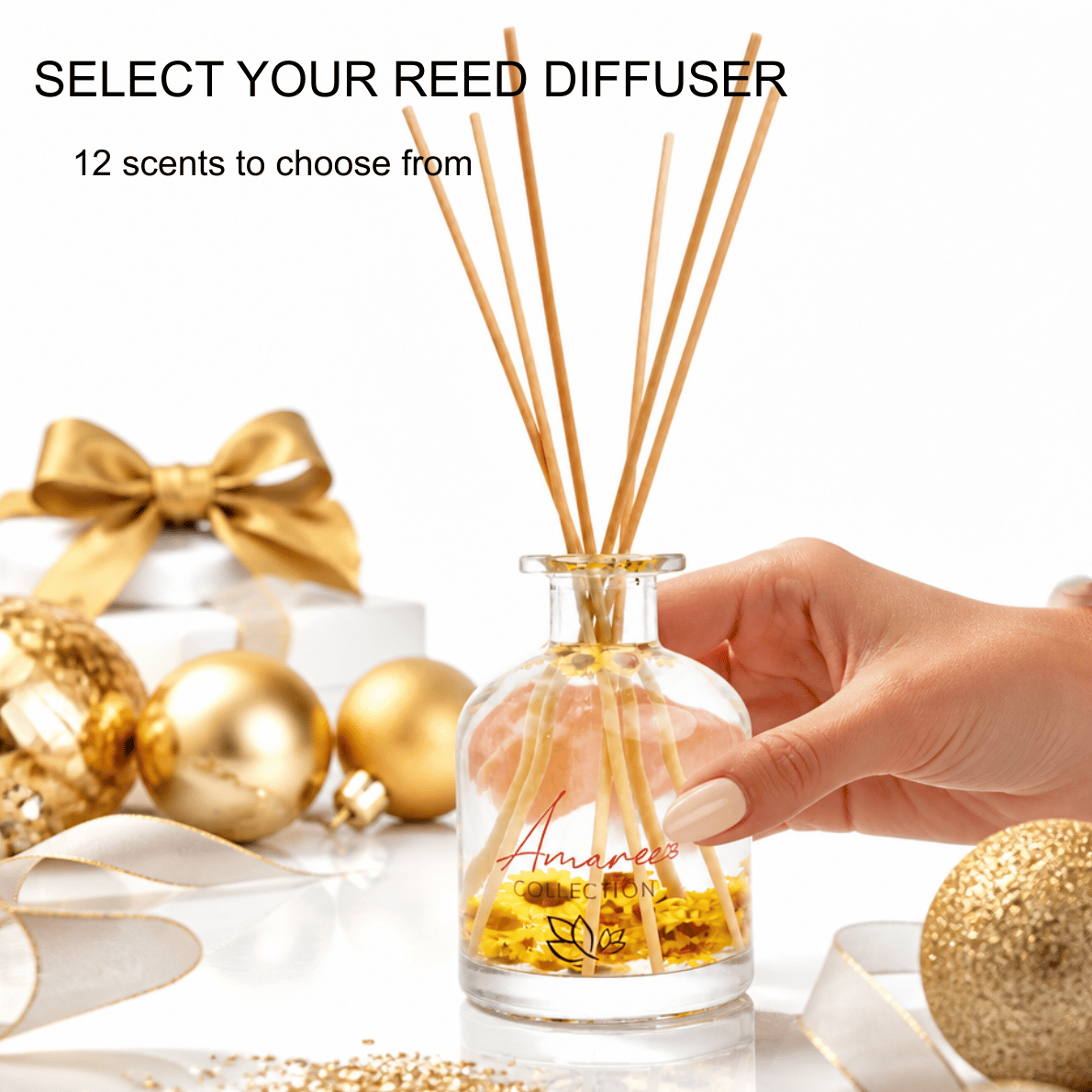 A clear glass reed diffuser bottle with natural wood reeds and yellow liquid, held by a hand. The scene includes gold holiday ornaments, a gold ribbon bow, and text reading &