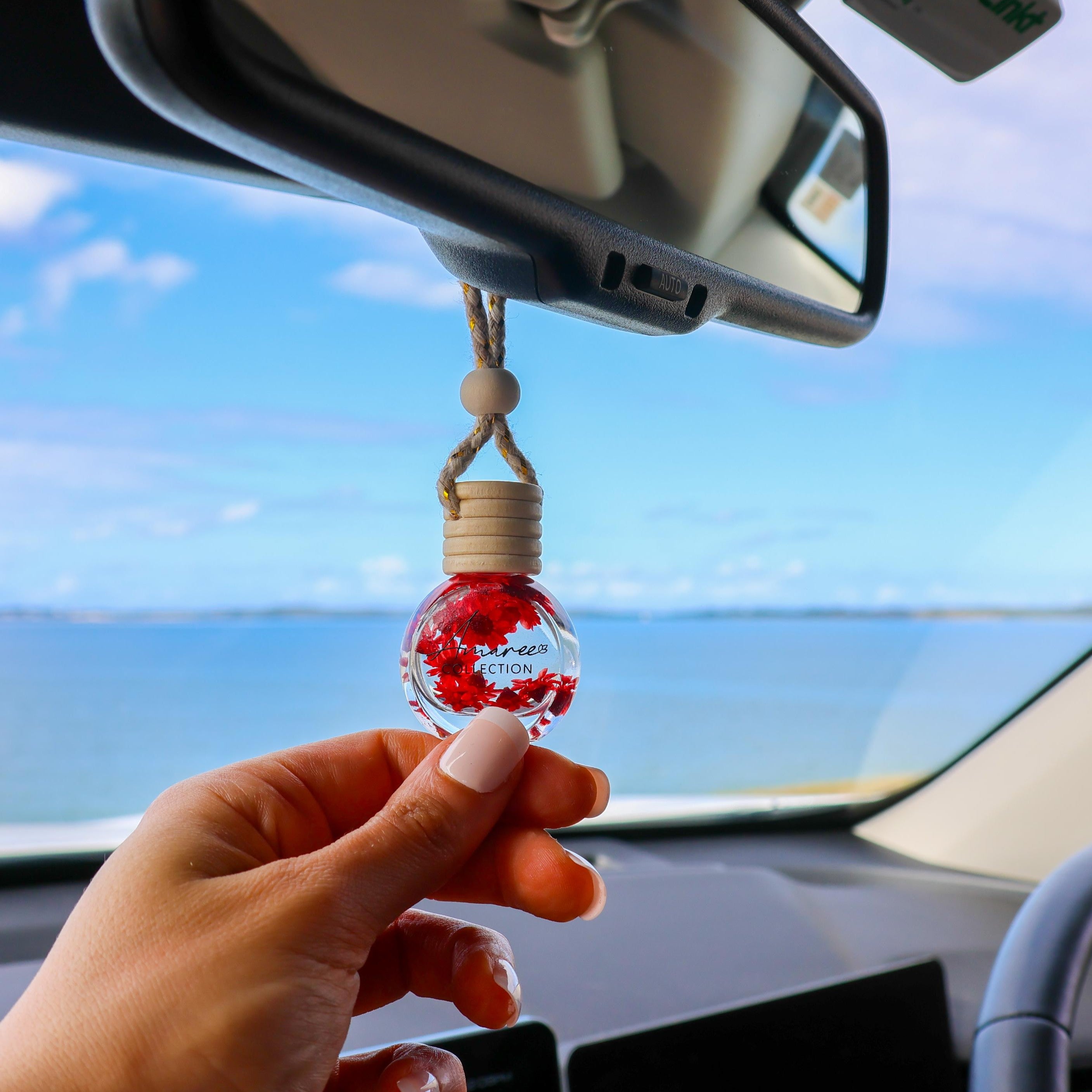 Car hanging ornament with red interior held by a hand inside a car with ocean view.