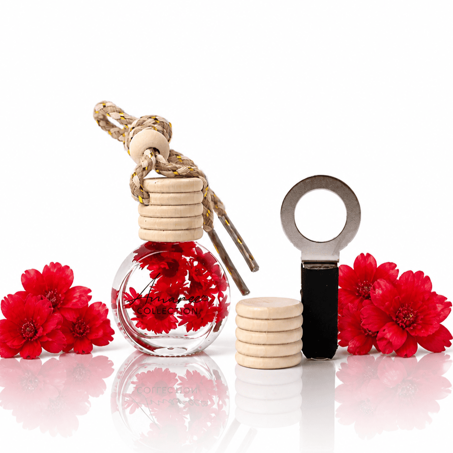 A decorative hanging car diffuser glass bottle with a cork stopper and hanging cord, filled with red flowers, shown beside a metal key ring and a stack of round wooden discs on a white reflective surface, with red flowers placed around it.