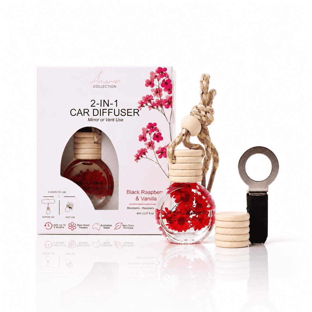 A 2-in-1 car diffuser set by Amara Collection, featuring a clear glass bottle filled with red liquid and wooden diffuser stones, alongside its white packaging box with pink raspberry illustrations. A metal clip and a stack of wooden diffuser stones are also shown on a reflective white surface.