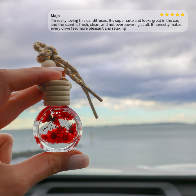 A hand holds a small glass car diffuser bottle with red maple leaves inside and a knotted cord, photographed in front of a cloudy sky and water. A review text bubble with a five-star rating appears at the top.