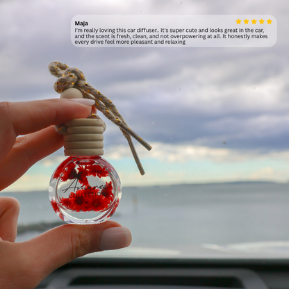 A hand holds a small glass car diffuser bottle with red maple leaves inside and a knotted cord, photographed in front of a cloudy sky and water. A review text bubble with a five-star rating appears at the top.