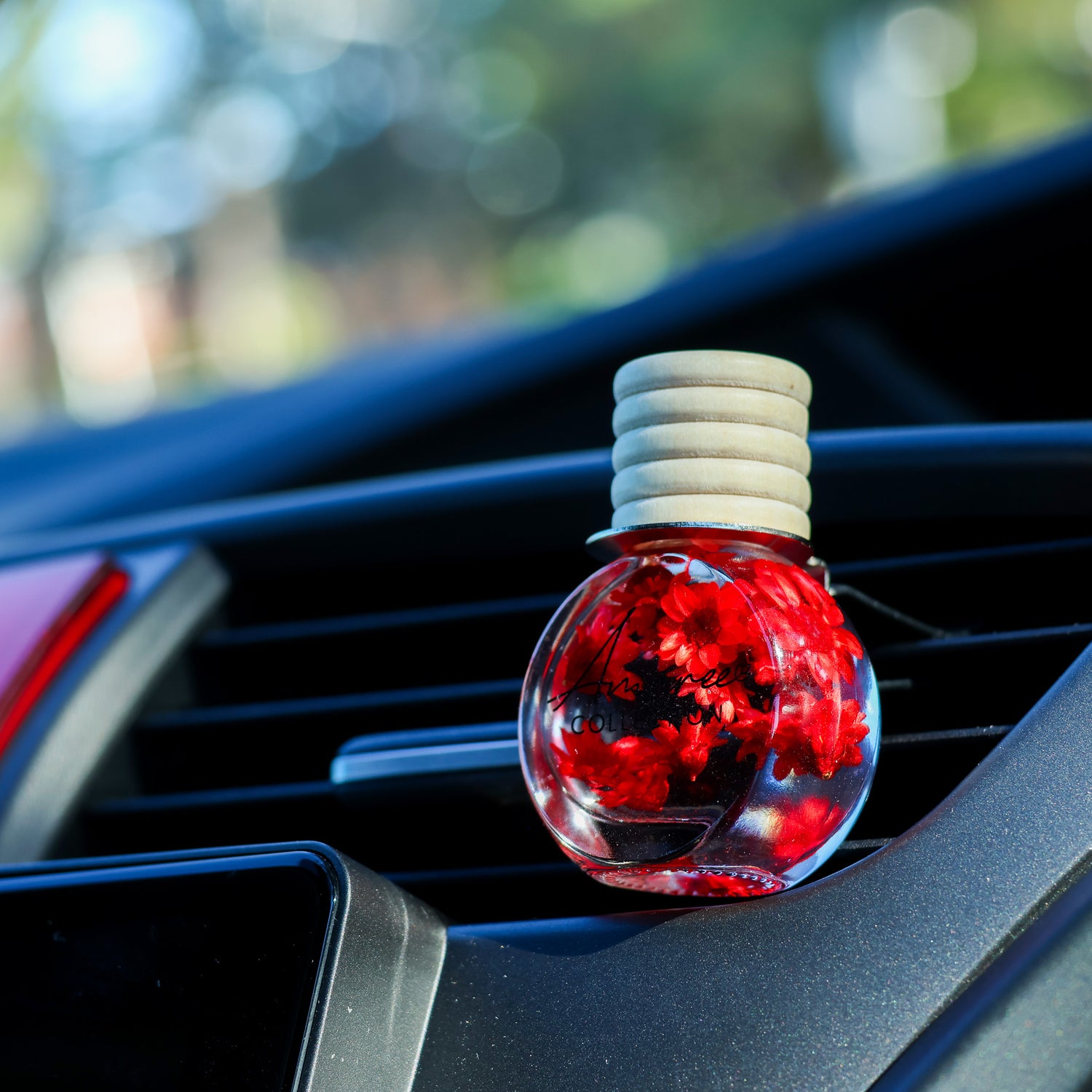 A round car air freshener bottle filled with red flowers and suspended in clear liquid, resting on a car dashboard vent with a blurred outdoor background.
