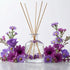 Japanese Honeysuckle Home fragrance diffuser bottle with wooden sticks surrounded by purple flowers on a white background