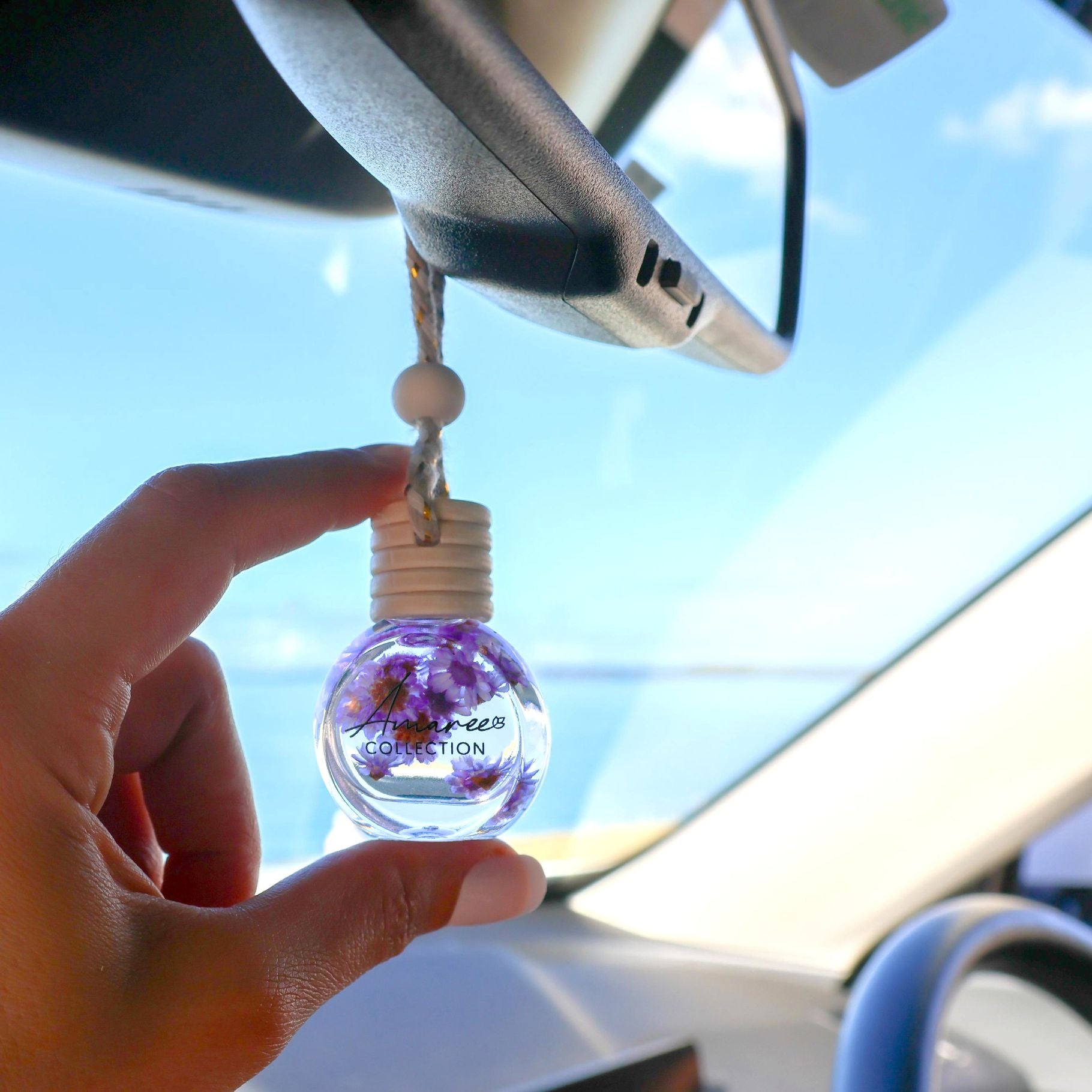 Hand holding a car air freshener with a clear bottle containing purple flowers, hanging from a car's rearview mirror.