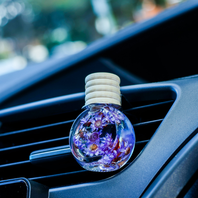A round car air freshener bottle with a light wood cap, filled with purple flowers and glitter, clipped onto a car dashboard air vent.