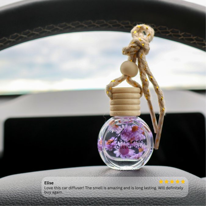 A car diffuser bottle with dried purple flowers inside a clear glass bottle, hanging from a knotted rope with wooden beads and a wooden cap, displayed on a car dashboard.