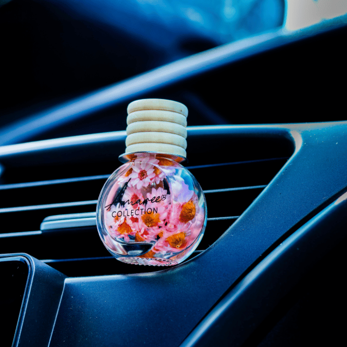 A round glass car air freshener bottle with dried orange and pink flowers inside, resting on a car air vent. The bottle has a light beige ribbed cap and the text &