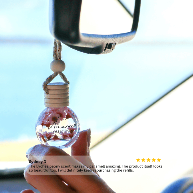 A hand holds a small clear glass car fragrance diffuser bottle with pink floral pieces inside, suspended from a rope and bead, near a car rearview mirror. The background is a bright, blurred car interior.