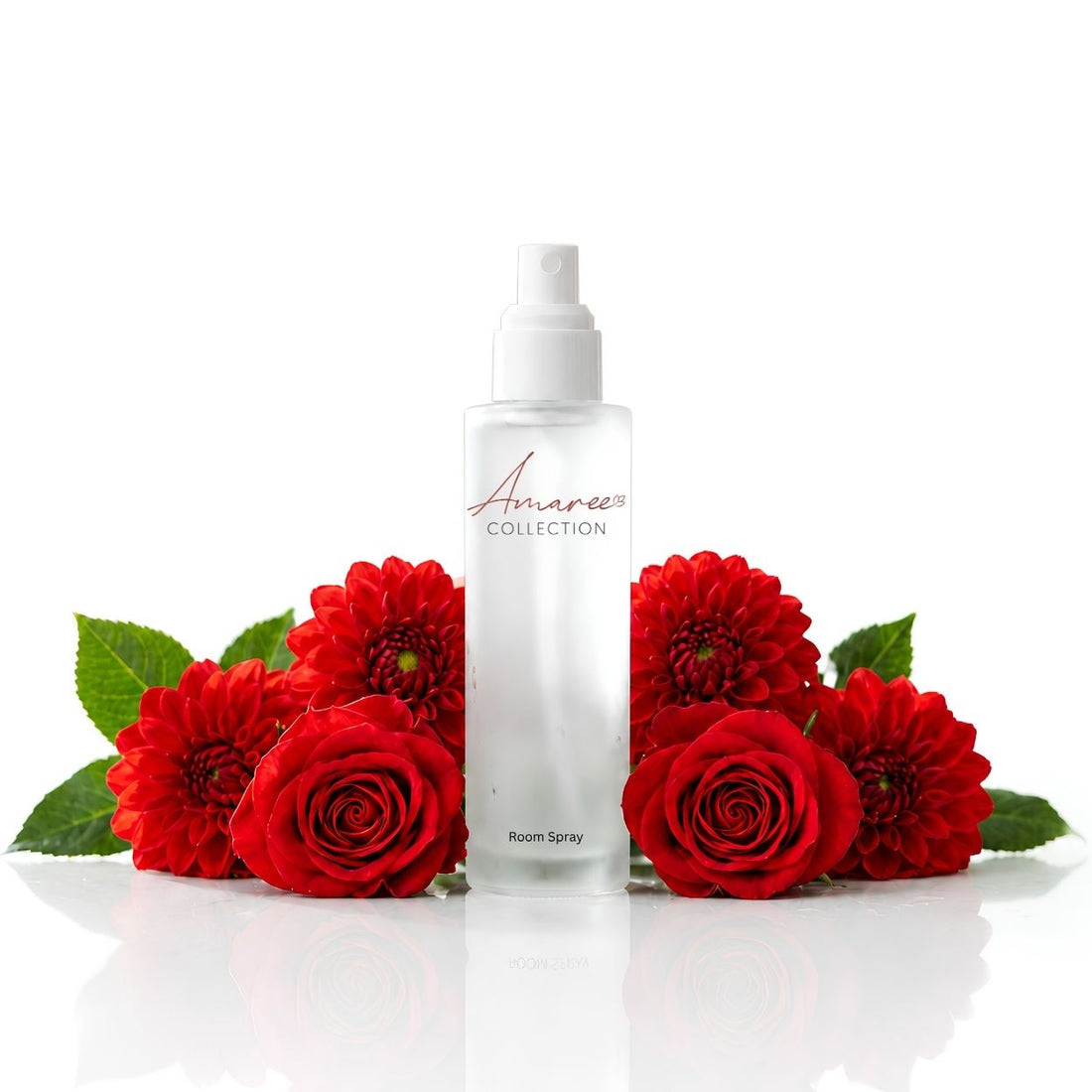 A clear room spray bottle from Amaree Collection, surrounded by vibrant red roses and green leaves on a reflective white surface against a white background.