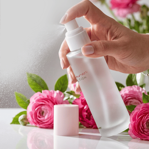 A hand holds a frosted white spray bottle, spraying a fine mist, with a small pale pink cap standing below it. Pink flowers and green leaves decorate the scene against a soft white background.