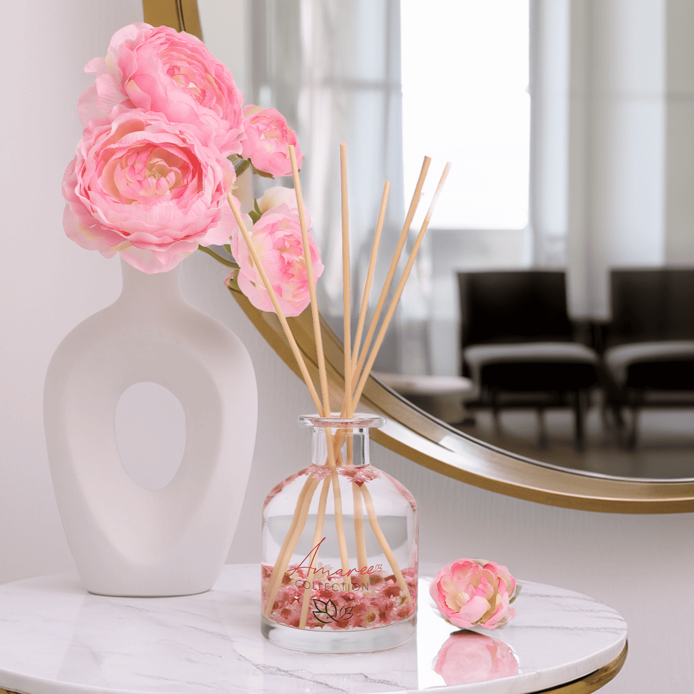 Decorative diffuser with pink flowers and sticks on a marble surface, with a blurred background of chairs and a mirror.