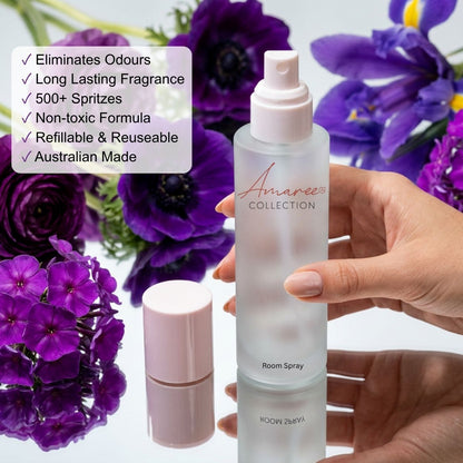 A hand holds a frosted glass room spray bottle with a pale pink cap next to its detached cap, with a list of product benefits overlaid on the left and purple flowers in the background.