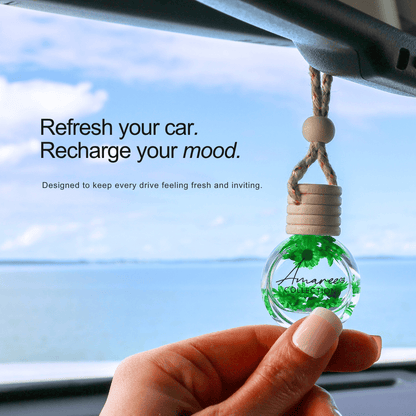 A hand holds a small clear glass car fragrance bottle with green leaves inside, hanging from a car&