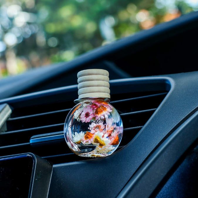 A clear round car air freshener bottle with dried multicolored flowers inside, resting on a black car dashboard vent with blurred greenery outside.