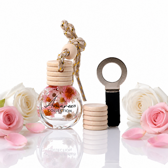 A clear glass diffuser bottle with a light wood cap and hanging rope, filled with dried flowers, shown beside a metal and black ring-shaped diffuser component on a white reflective surface. White and pink roses and petals are arranged around the products.