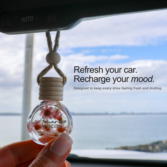 A hand holds a clear glass car air freshener bottle with dried flowers inside, hanging from a rope loop. The background shows a car interior and a scenic ocean view through the window, with promotional text on the right.