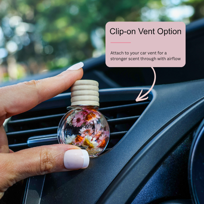 A hand with white manicured nails holds a small round car air freshener diffuser with a wooden cap and colorful dried flowers inside, positioned against a car vent. A pink text box reads &
