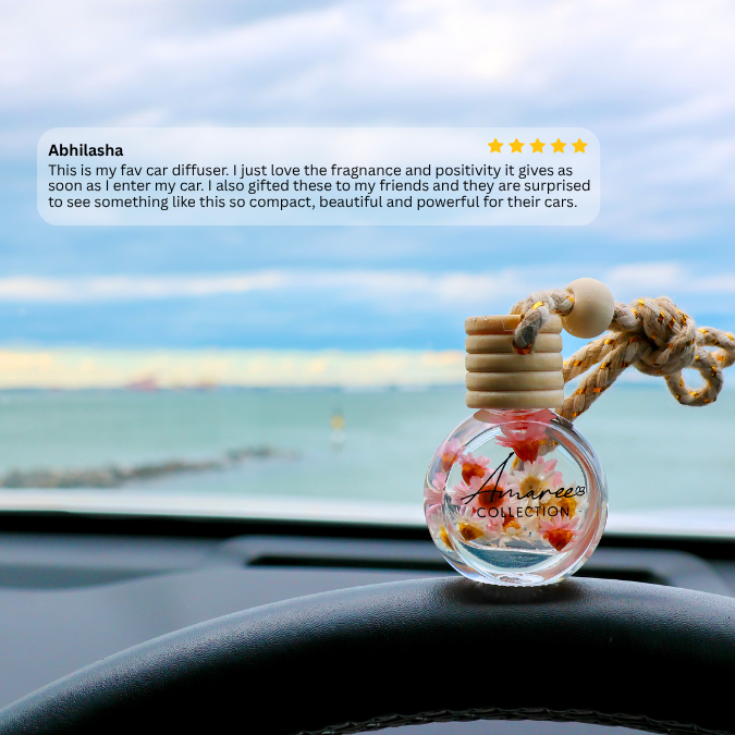 A clear glass car diffuser bottle with a wooden stopper and knotted rope, filled with colorful flower pieces, resting on a car dashboard with a blurred ocean view outside. A review text bubble appears on the left.