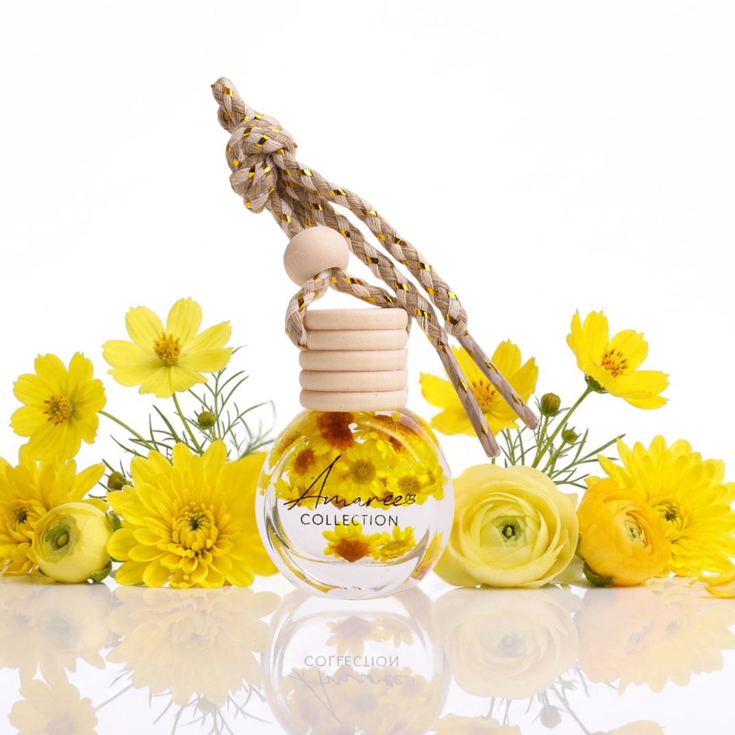 Car air freshener with wooden holder and clear bottle surrounded by yellow flowers on a reflective surface.