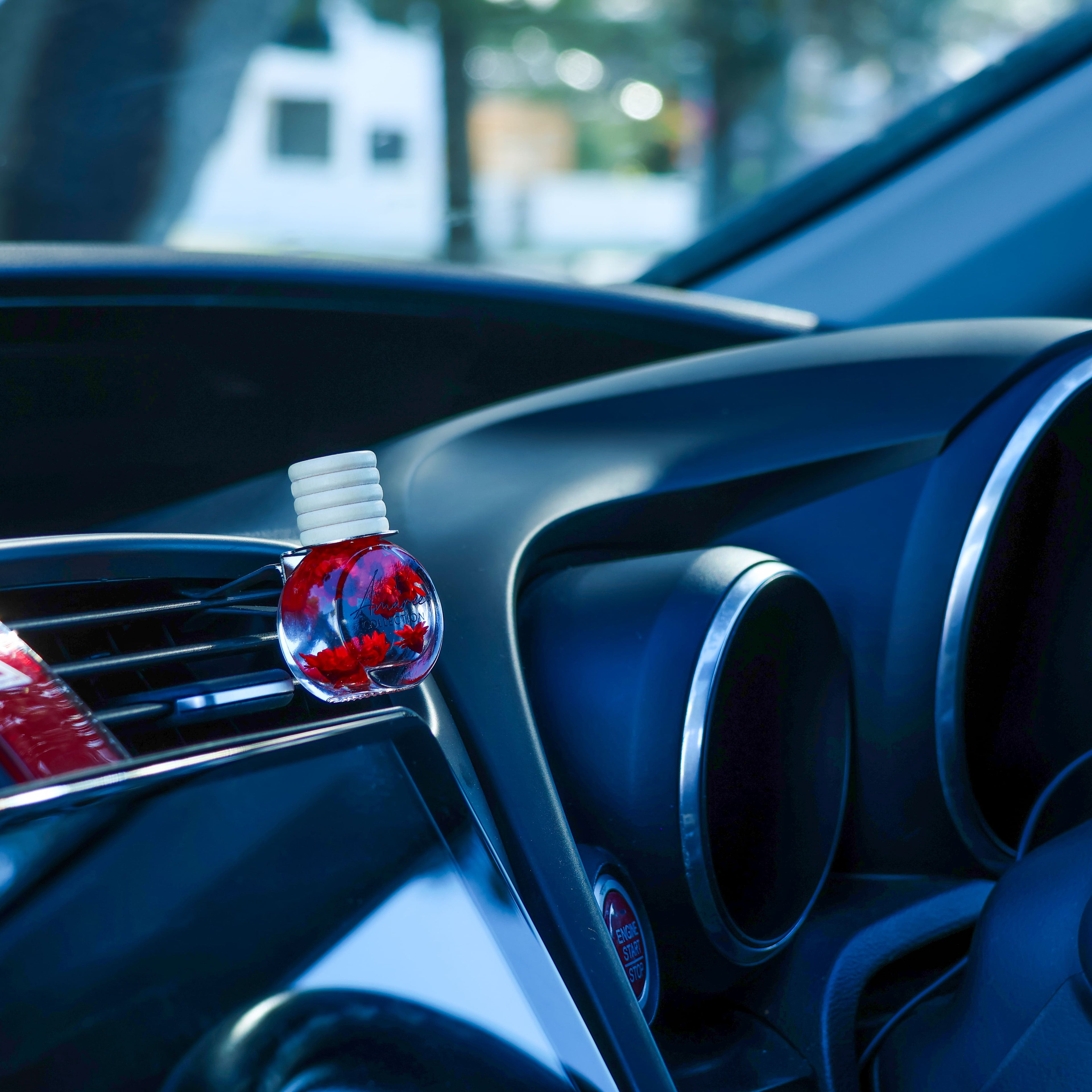 A clear round car air freshener dropper bottle with red liquid and a white cap, resting on the dashboard of a blue car.
