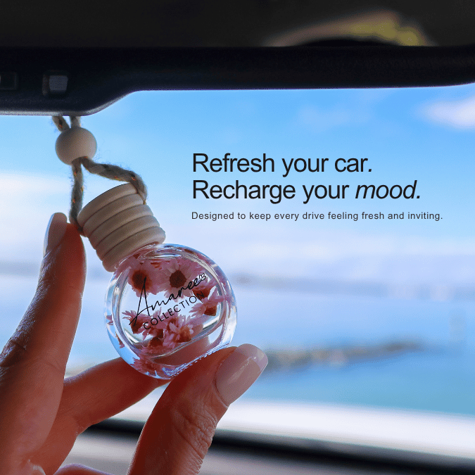 A hand holds a small clear glass car fragrance diffuser bottle with dried pink flowers inside and a beige ribbed cap, positioned in front of a car window showing a blurred blue sky and water. Text on the image reads, &