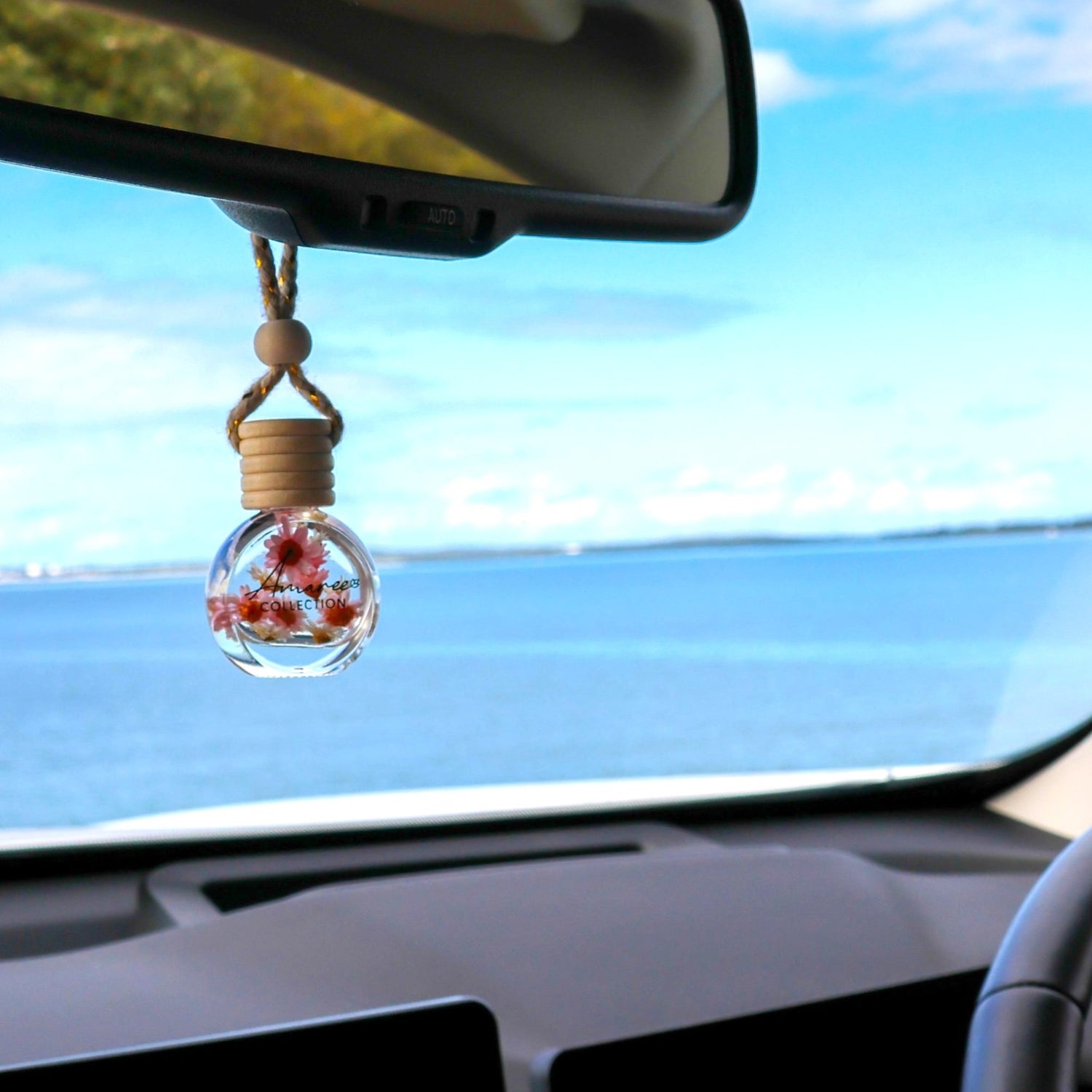 Car interior with a decorative hanging item featuring flowers, overlooking a scenic ocean view.